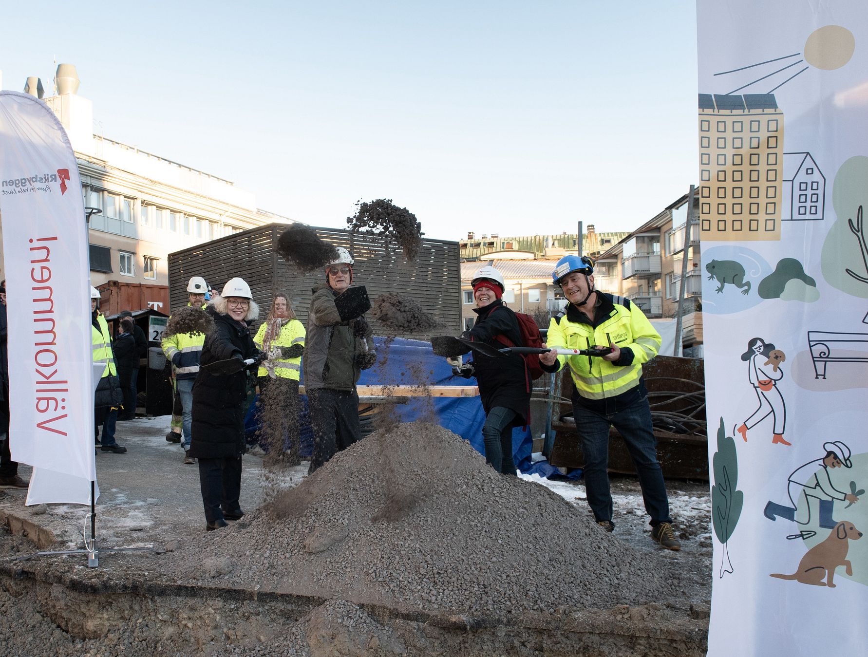 Människor i arbetskläder skyfflar grus på en byggarbetsplats, omgivna av banderoller och en stadsmiljö i bakgrunden