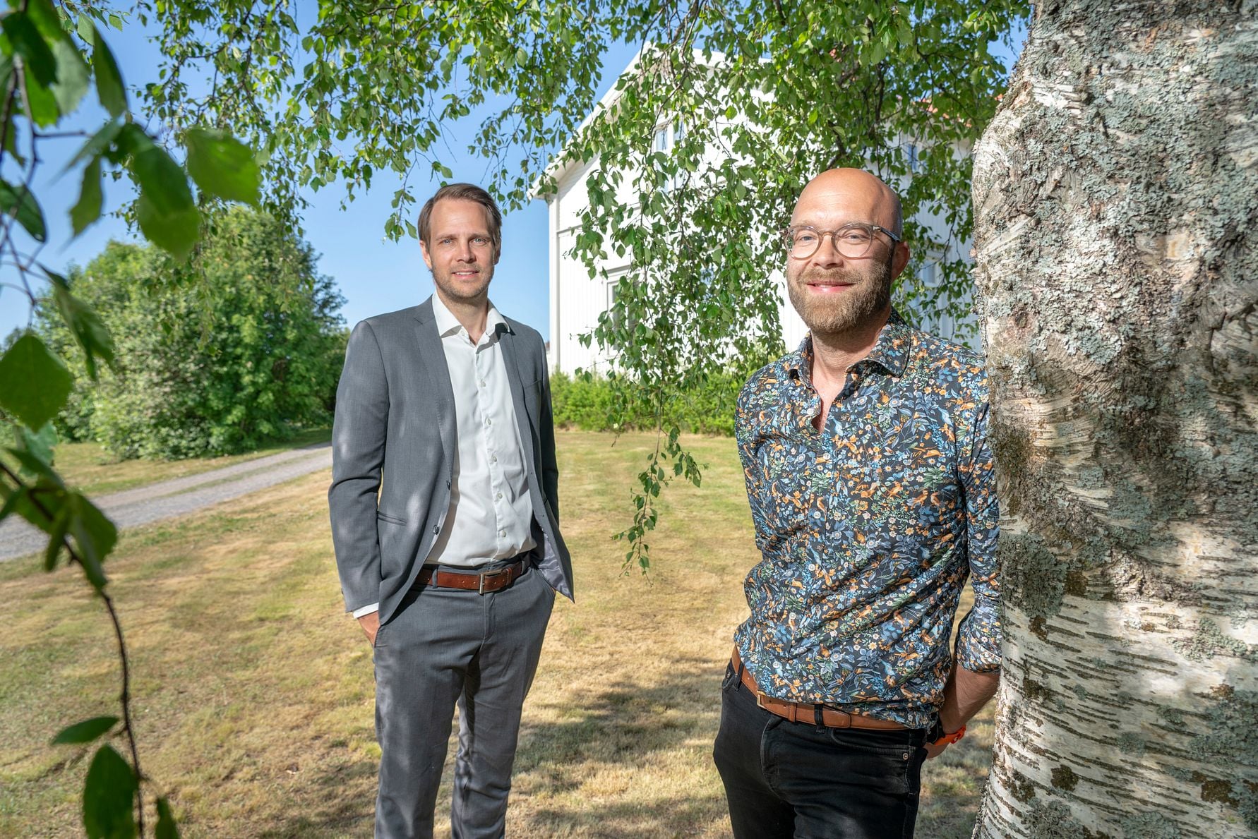 Två personer står utomhus på en gräsmatta nära ett träd, med en vit byggnad och träd i bakgrunden under en klarblå himmel
