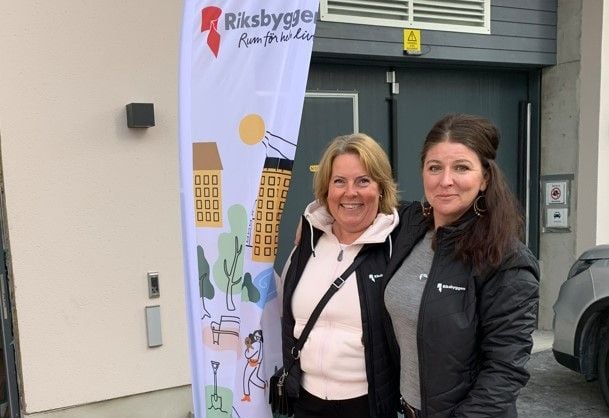 Två personer står framför en byggnad bredvid en flagga med Riksbyggens logotyp och texten "Rum för hela livet