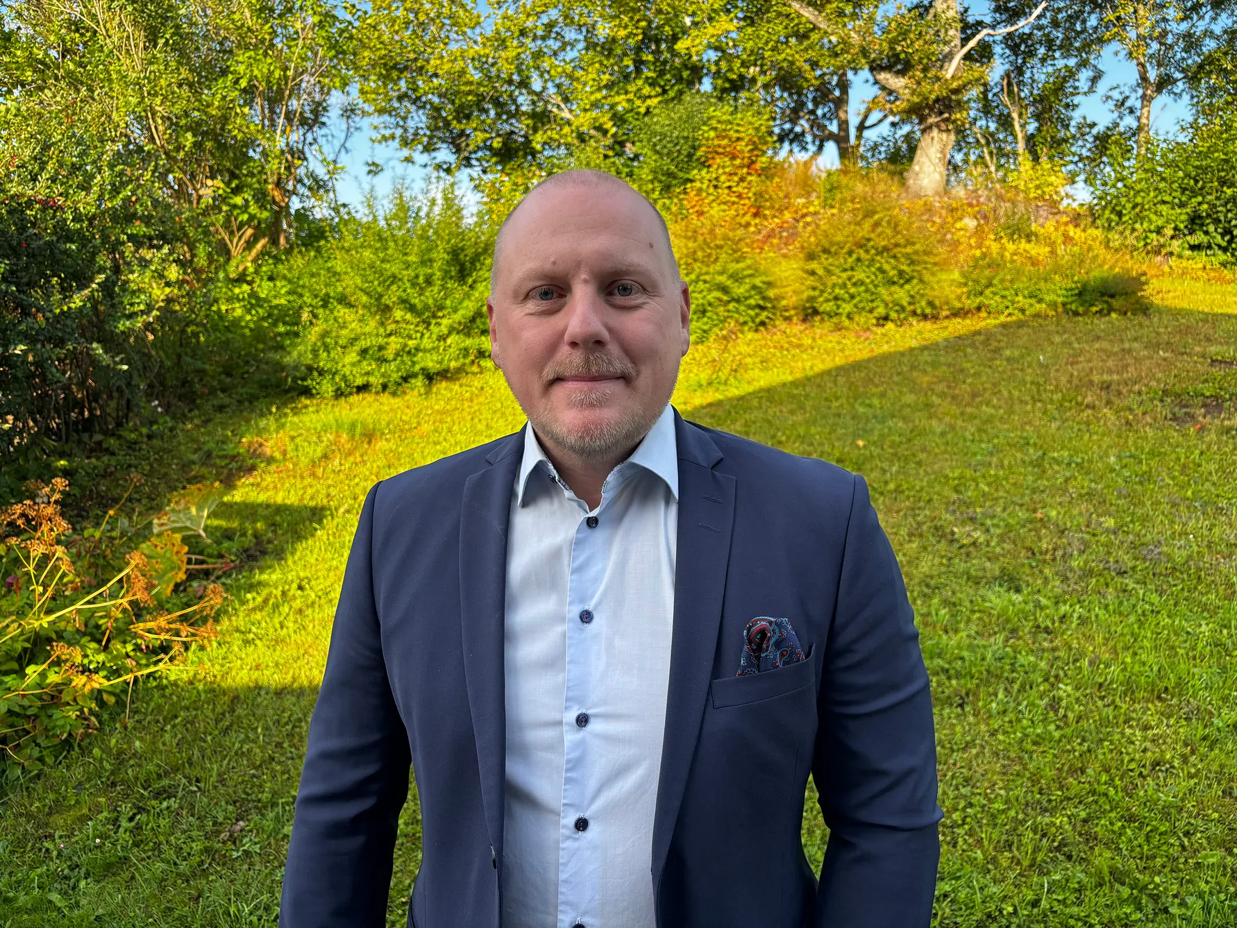 Man i blå kostym står utomhus i grönskande miljö