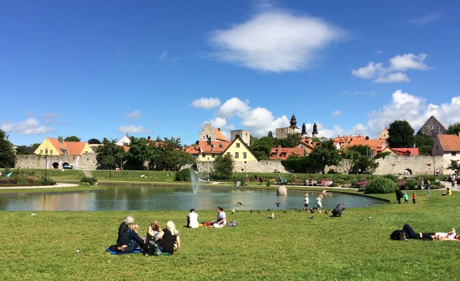 Grön park med människor som sitter på gräsmattan, en damm med fontän i mitten och historiska byggnader i bakgrunden under en blå himmel