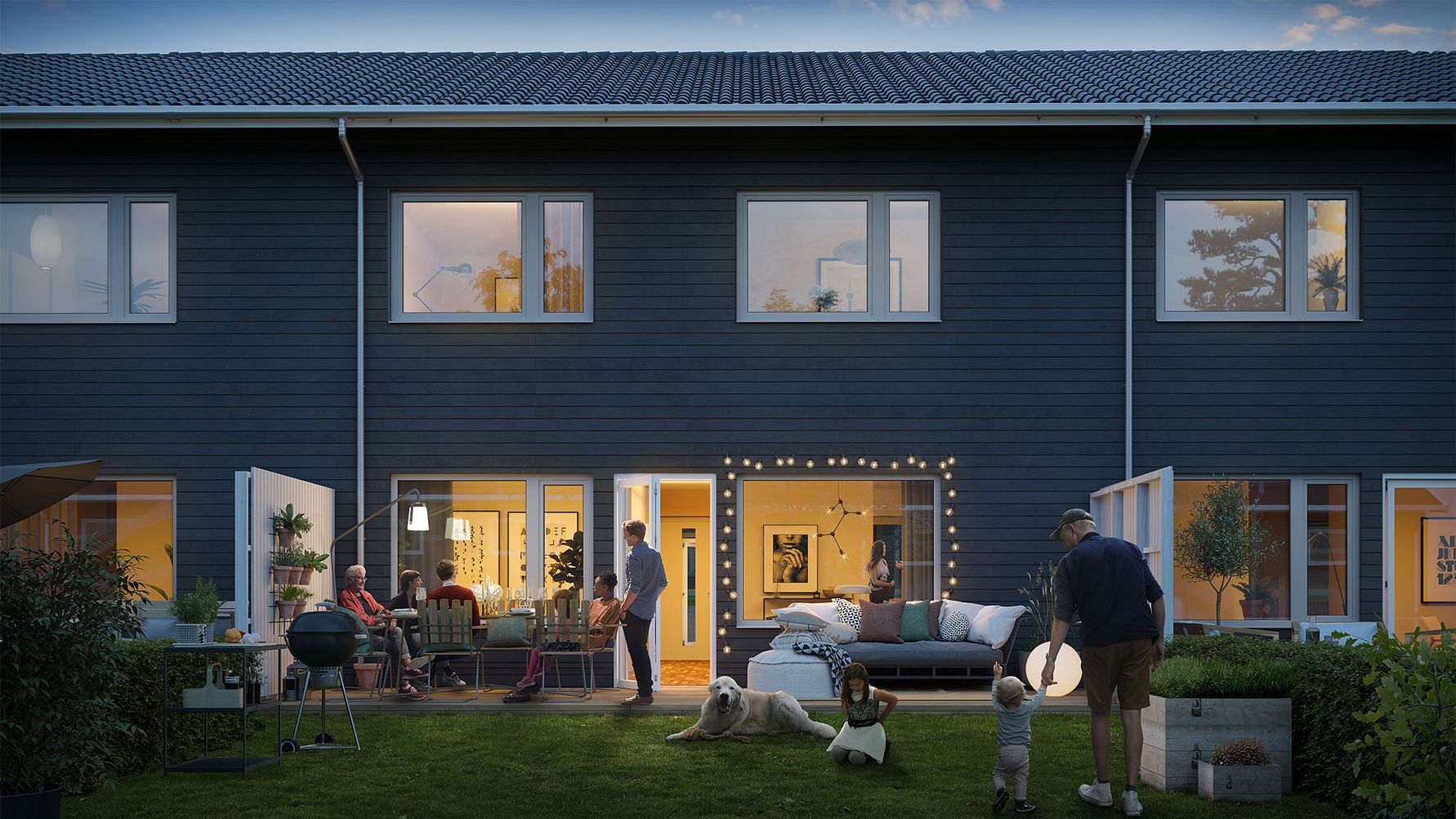 Tvåvåningshus med mörk fasad på kvällen, med människor som umgås i trädgården och interiören upplyst