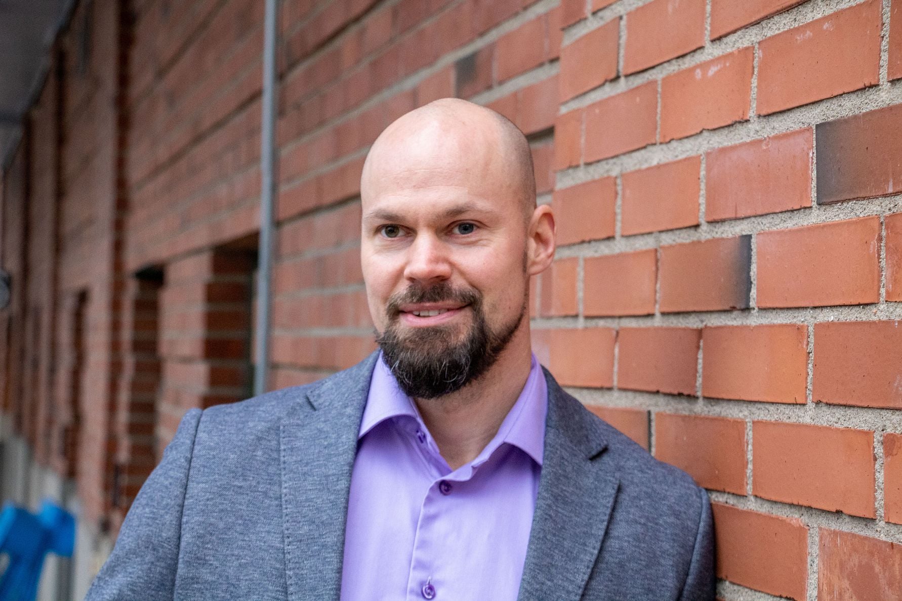 Man i ljuslila skjorta och grå kavaj lutad mot en tegelvägg