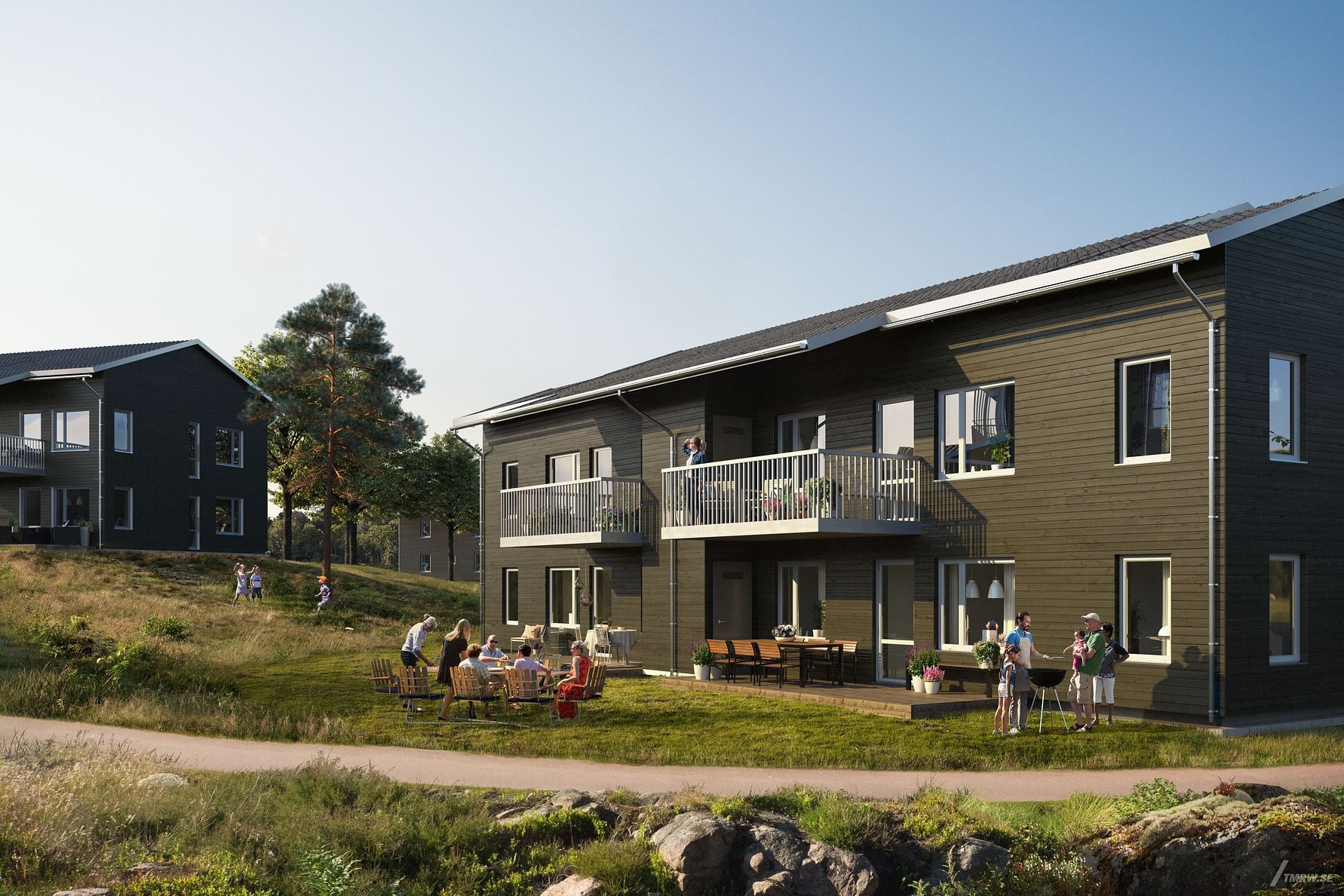 Två moderna bostadshus med mörk träfasad på en grönskande plats, personer samlade vid ett bord och barn som leker utomhus i dagsljus