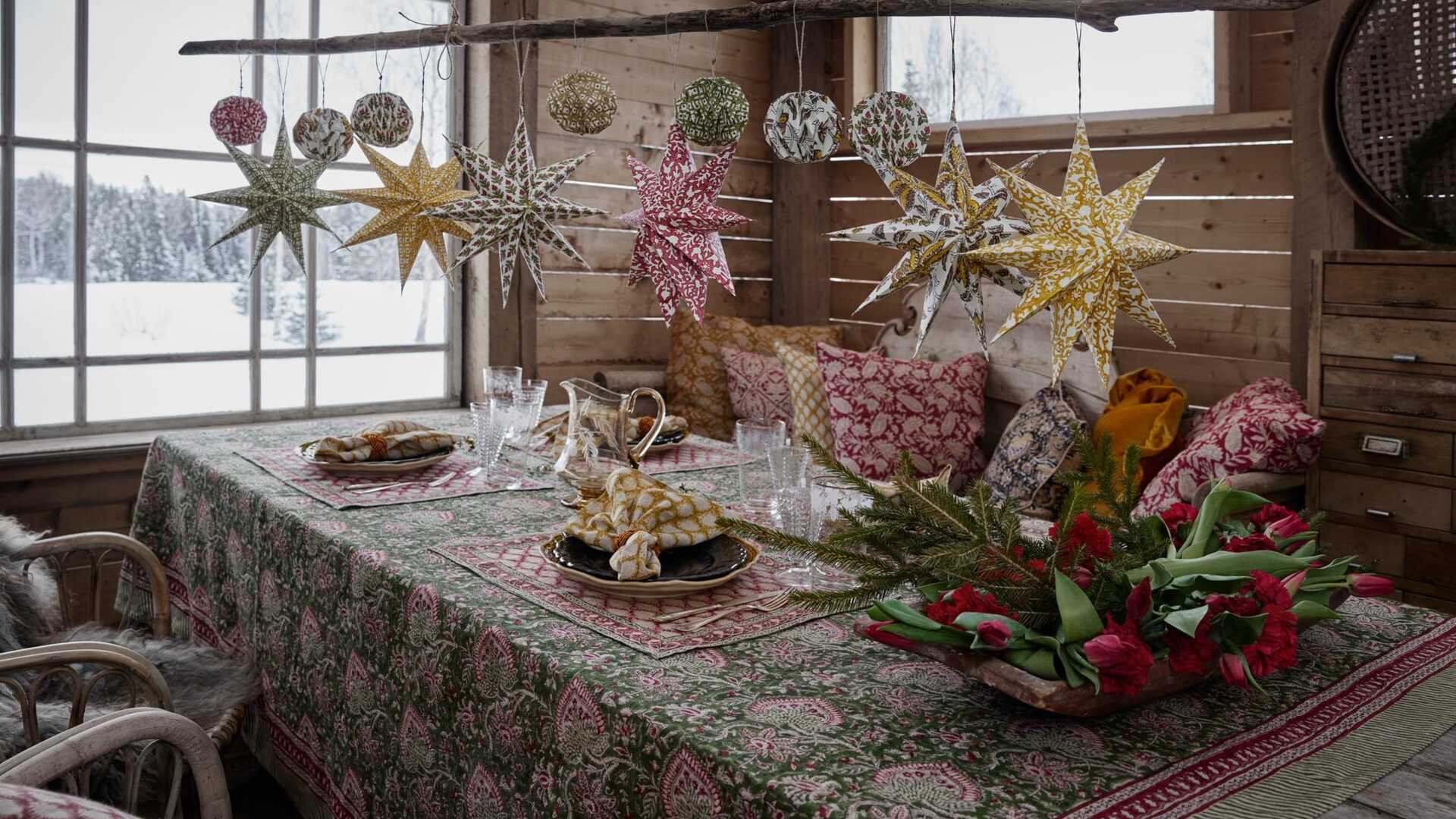Rustik matplats inredd med mönstrade stjärnor och ornament, med blommor och tallkvistar på ett dukat bord med utsikt över en snöig skog