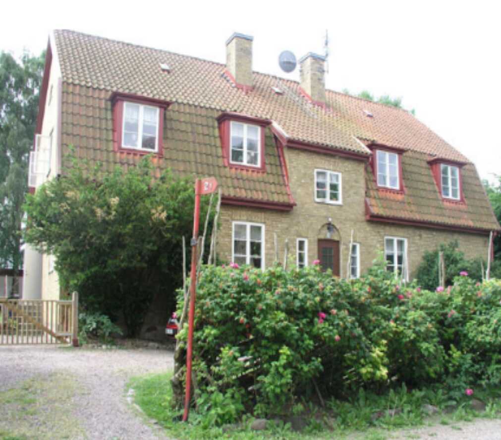 Tegelhus med brunt tak och vita fönster omringat av grönska och en grusgång