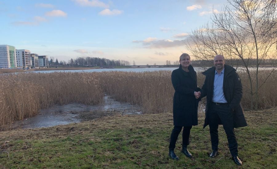 Två personer står på en gräsmatta nära en sjö med vass och omgivande träd, medan de skakar hand. I bakgrunden syns flerfamiljshus och en klar himmel med moln