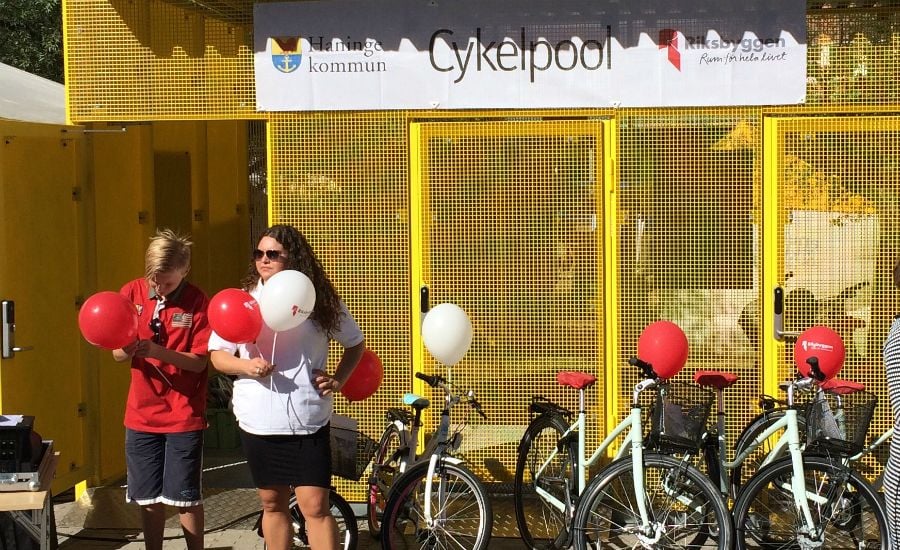 En cykelpool med gula metallväggar i bakgrunden, flera uppställda cyklar och två personer som håller i röda och vita ballonger