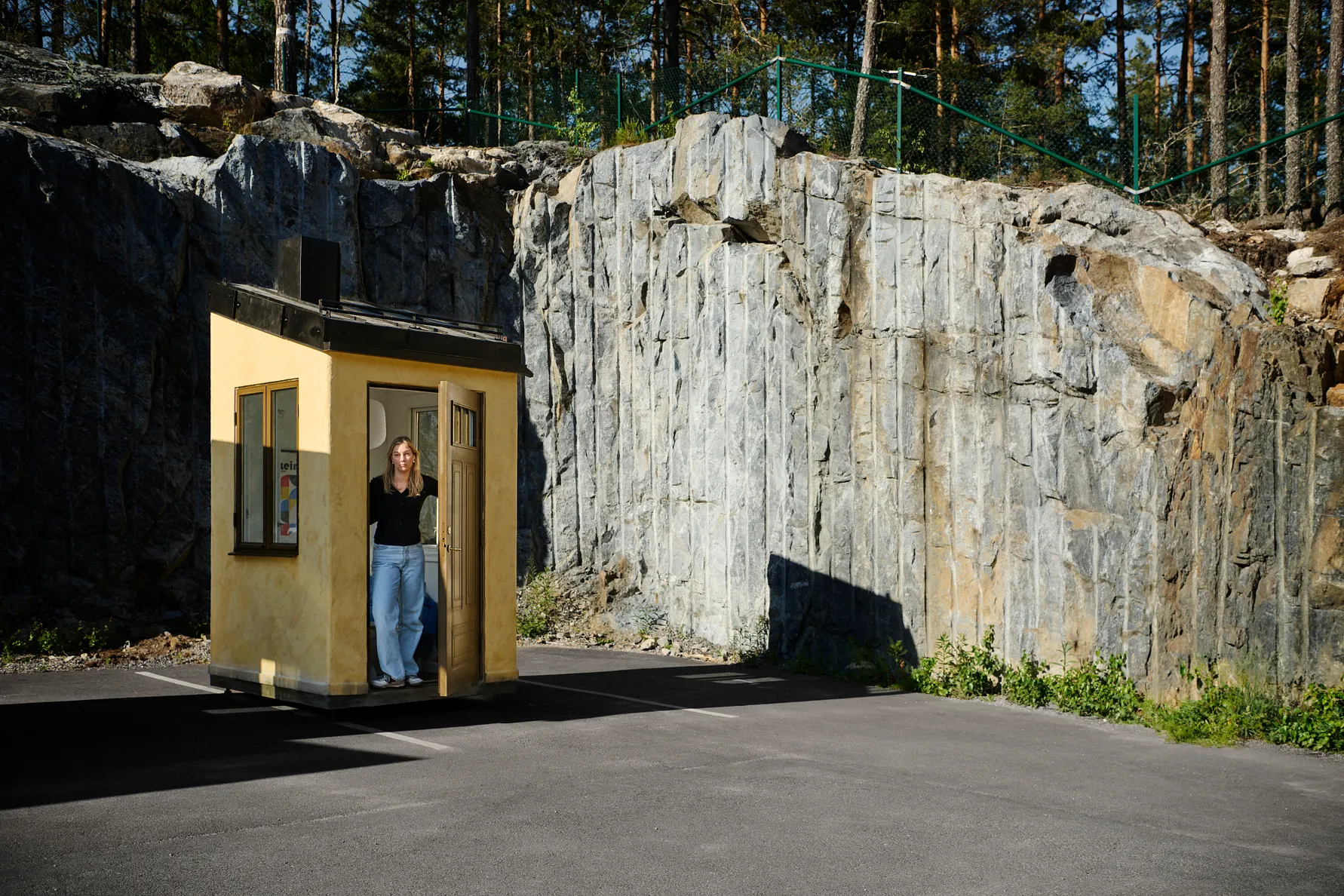 Person står i en liten gul byggnad vid en bergvägg, med skog och asfalt runt omkring