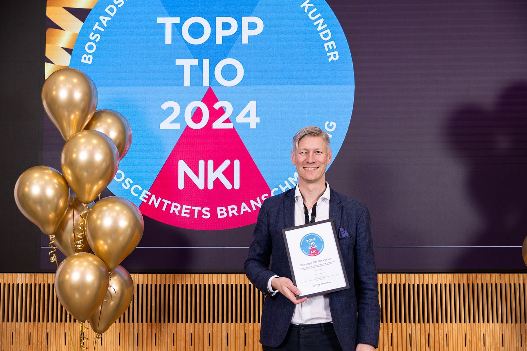 Man i mörk kavaj håller ett diplom framför en skärm med texten "TOPP TIO 2024 NKI" och omgivande guldfärgade ballonger