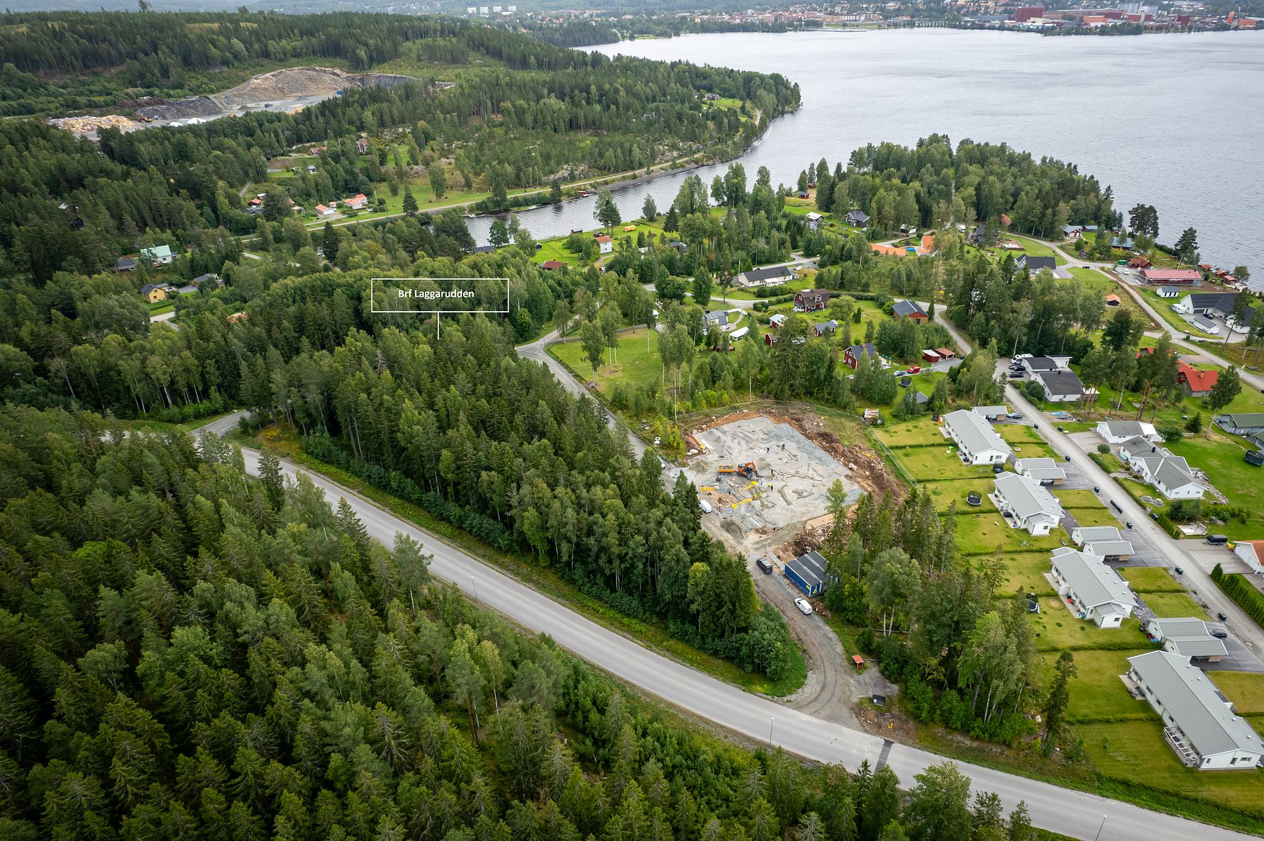 Flygbild av ett bostadsområde omgivet av skog, med en väg som slingrar sig genom landskapet och en sjö i bakgrunden