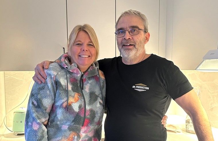 Två personer står i ett kök, varav en bär en mönstrad tröja och den andra en svart t-shirt med tryck. Bakgrunden visar ljusa köksskåp och en bänk med föremål