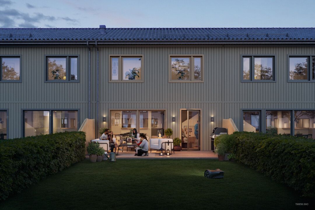 Radhus med träfasad och flera fönster, omgiven av grönska och upplyst trädgård under kvällstid