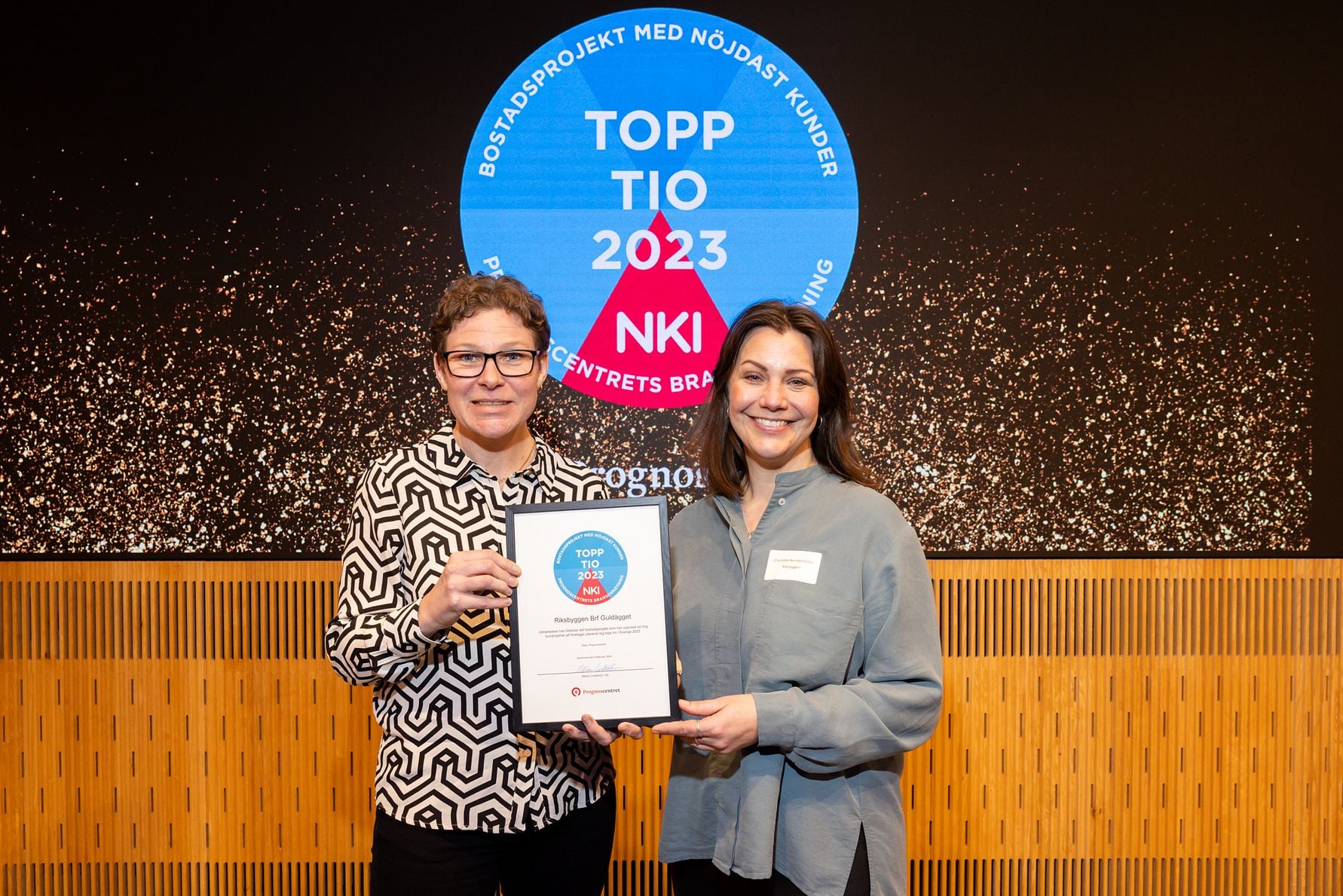 Två personer står framför en skylt med texten "Topp Tio 2023 NKI" och håller upp ett diplom