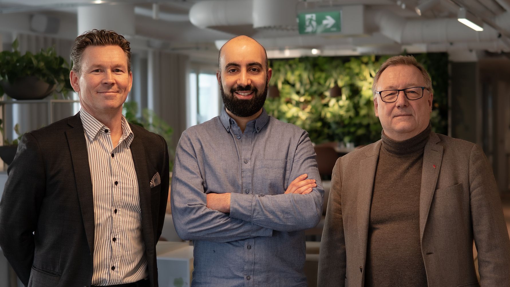 Tre personer står i ett modernt kontorsutrymme med växter och ljusa väggar i bakgrunden