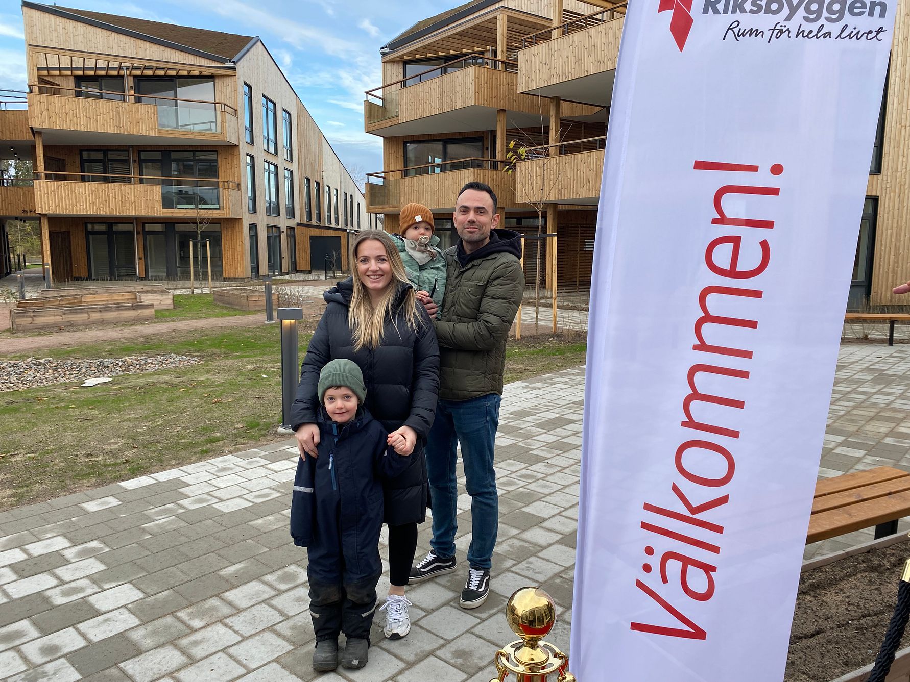 En familj står utomhus på en stenlagd gång framför moderna trähus och en flagga med texten "Välkommen!