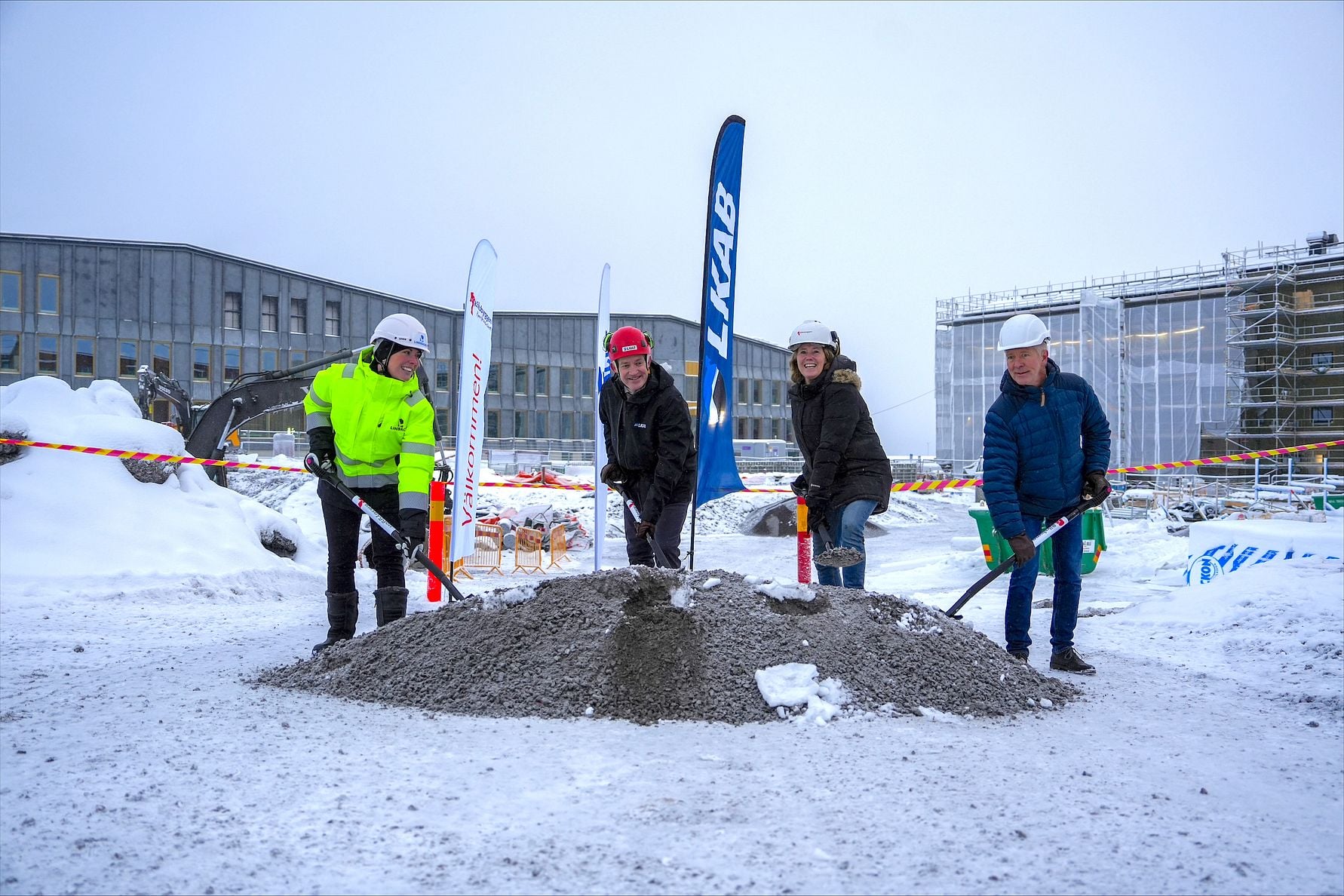 Fyra personer iförda skyddsutrustning står runt en hög med grus på en snöig byggarbetsplats, med byggnader och företagsflaggor i bakgrunden