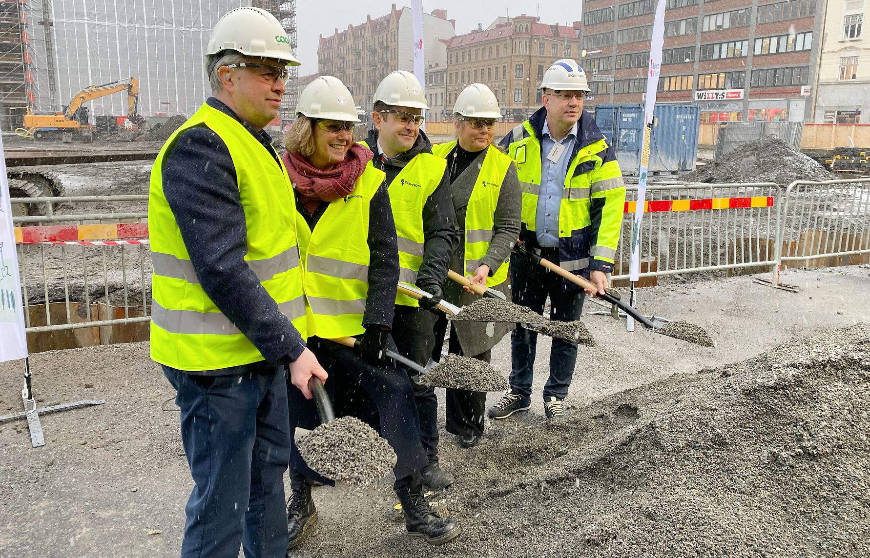 Personer i byggarbetskläder och reflexvästar håller i spadar framför en grushög på en byggarbetsplats. Bakgrunden visar byggnader, byggutrustning och säkerhetsavspärrningar