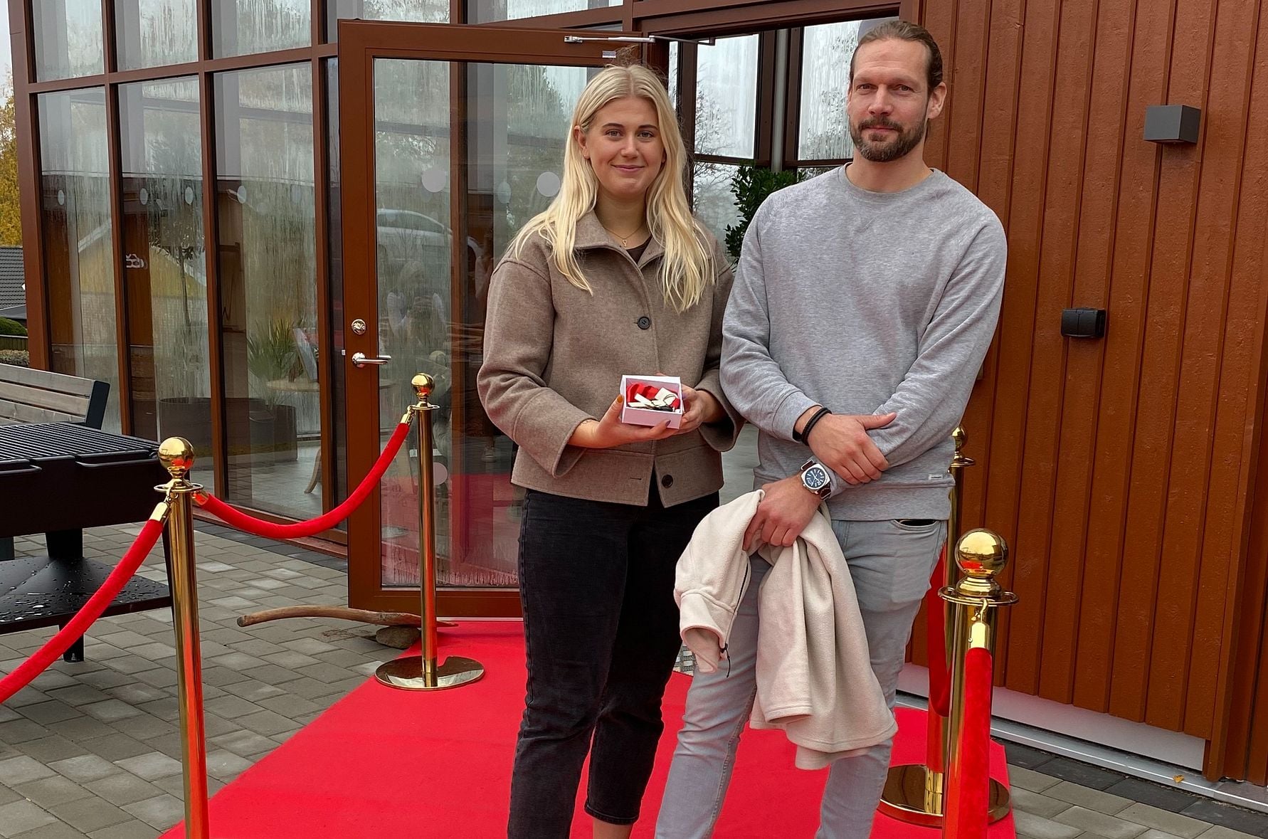 Två personer står på en röd matta framför en byggnad med glasdörrar, en av dem håller en liten låda med blommor