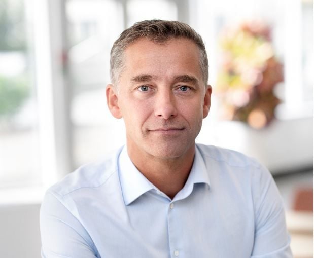 Man i ljusblå skjorta sitter inomhus med suddig bakgrund som visar växter och ett stort fönster