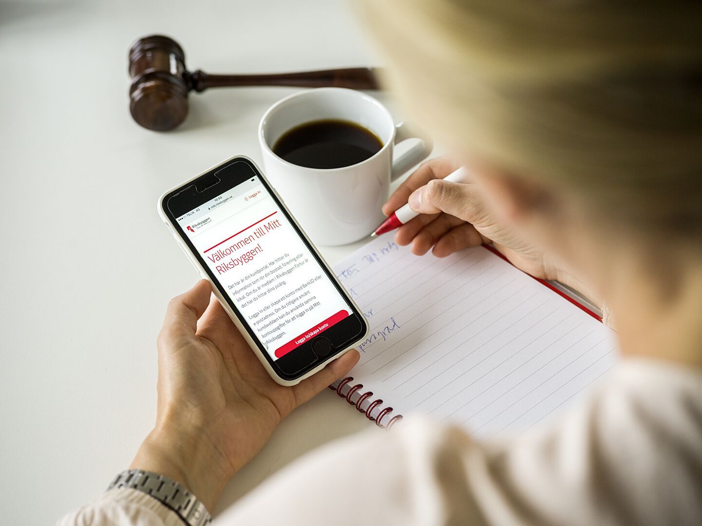 En person håller en mobiltelefon med en webbplats synlig, bredvid en kaffekopp, anteckningsblock och penna på ett bord