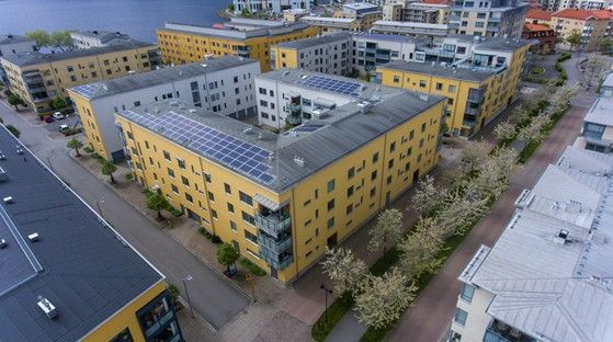Flygbild över ett bostadsområde med gulmålade flerfamiljshus, solpaneler på taket och blommande träd längs gångvägen