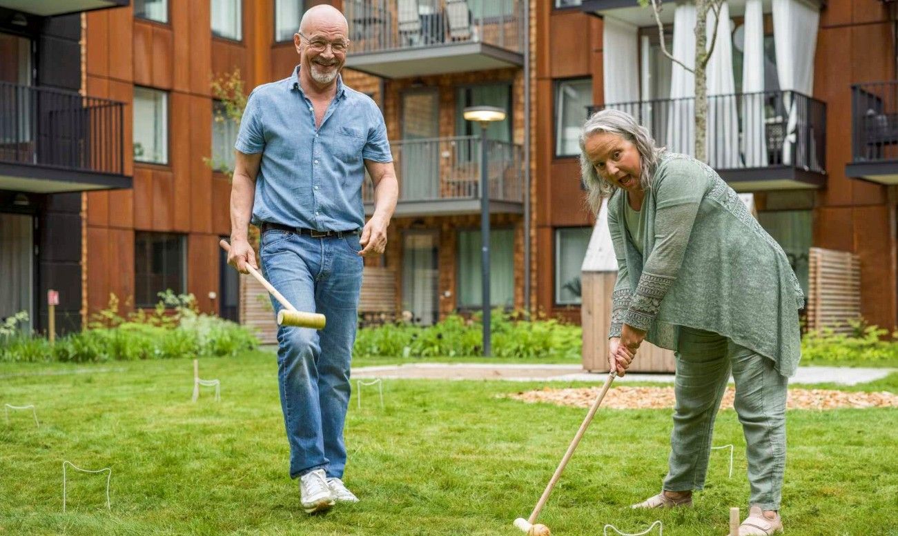 Två personer spelar krockett på en gräsmatta framför ett modernt bostadshus med balkonger