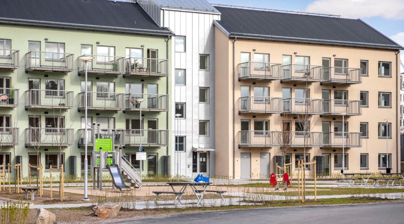 Flerbostadshus med färgade fasader i grönt och beige, balkonger, lekplats med gungor och grönområde i förgrunden