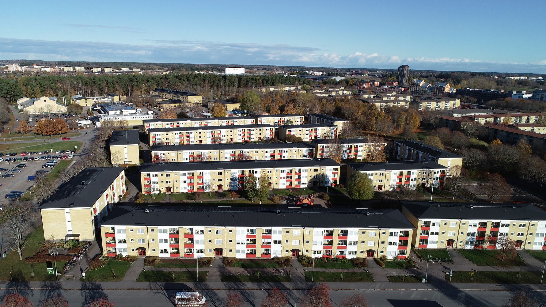 Flerbostadshus med gul fasad och röda detaljer, omgivna av träd och öppna ytor i en förortsmiljö under en klar himmel