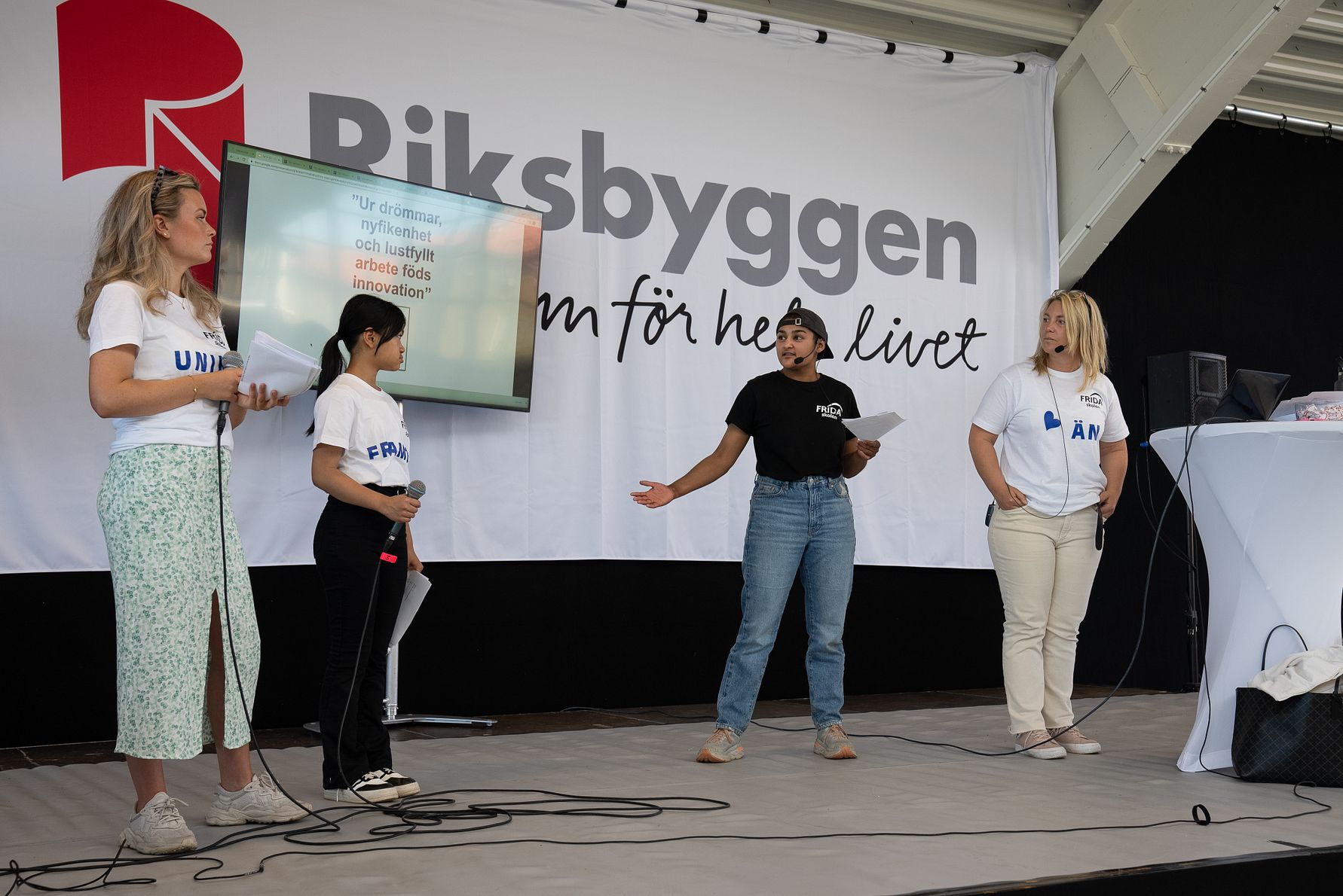 Fyra personer står på en scen framför en Riksbyggen-banner, med en presentation på en skärm i bakgrunden