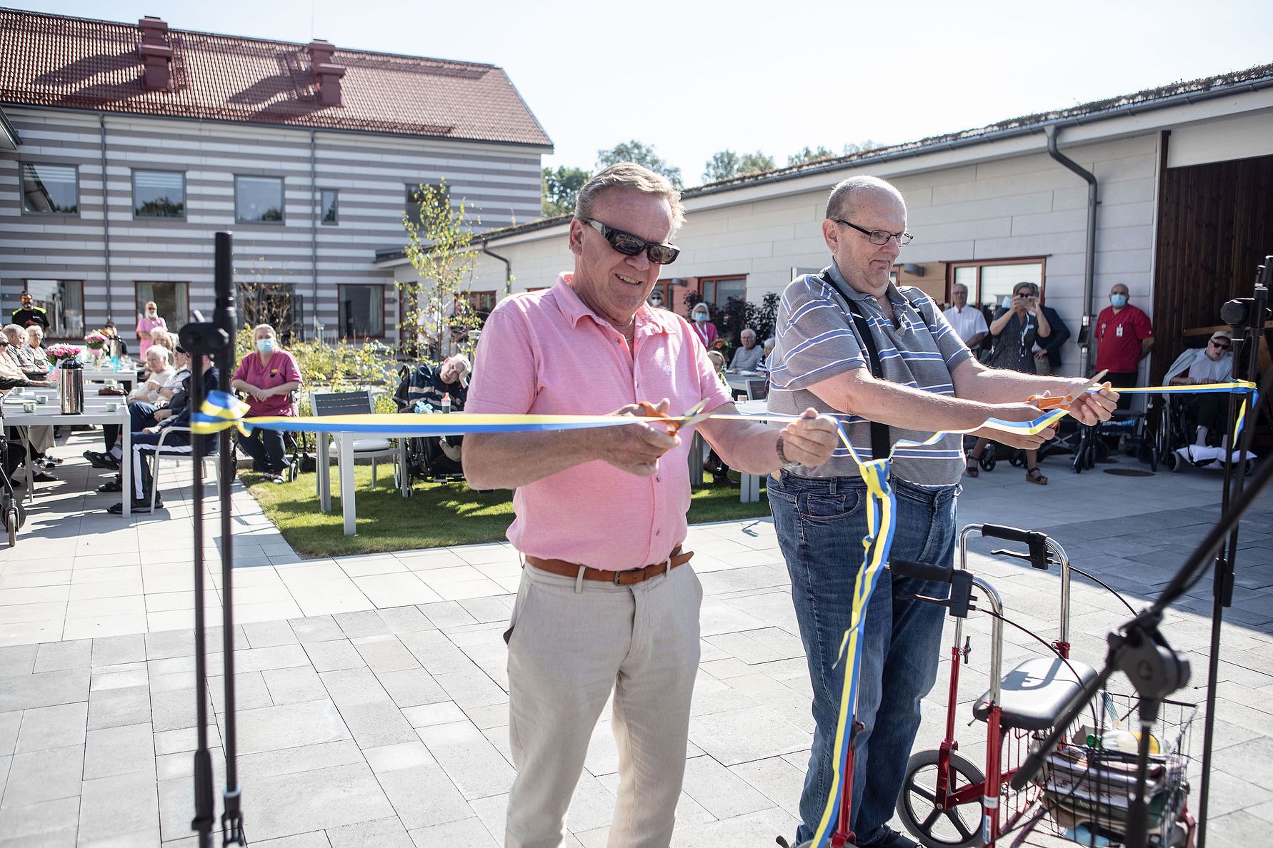 Två personer håller i ett band under en bandklippningsceremoni utomhus, omgivna av människor och byggnader i bakgrunden
