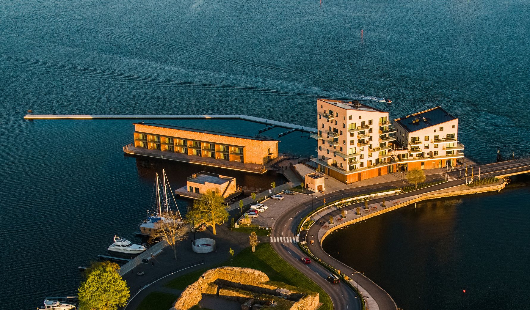 Luftvy över en modern byggnadskomplex vid vattnet med båtar, bryggor och en väg som slingrar runt området