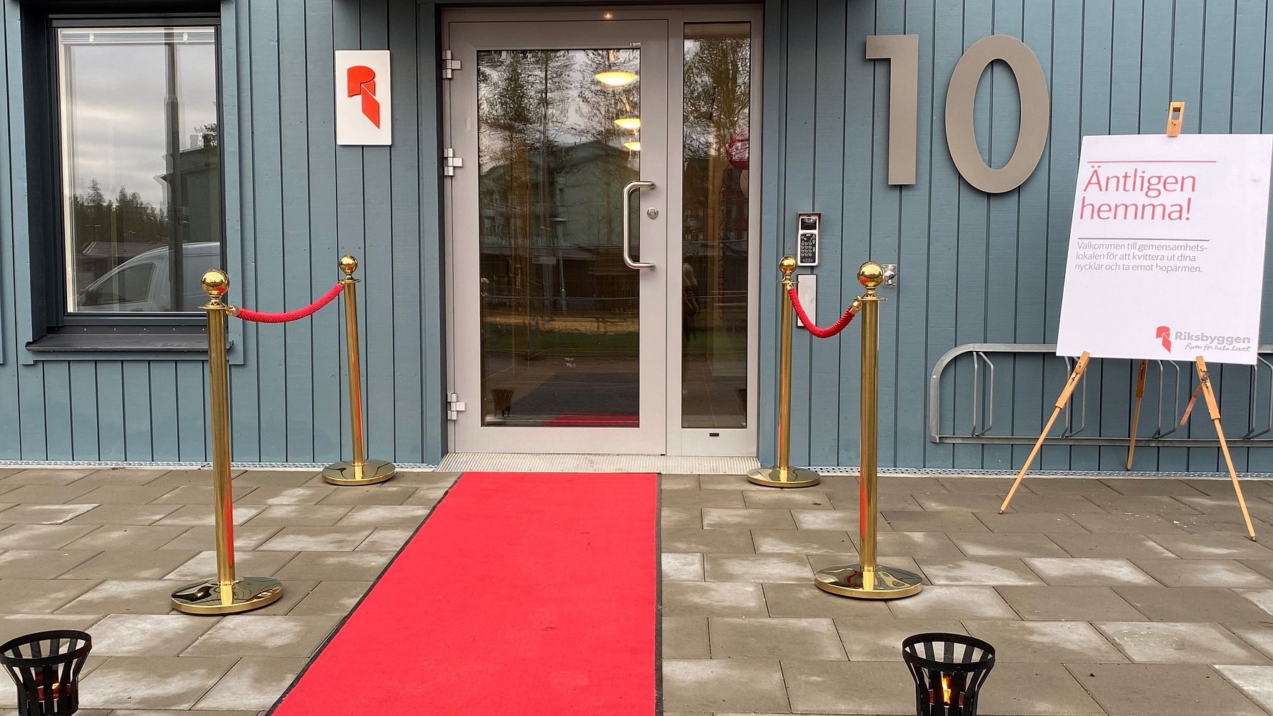 Ingång till en byggnad med blå fasad, röd matta, och avspärrningsstolpar. En skylt på ett staffli står bredvid dörren
