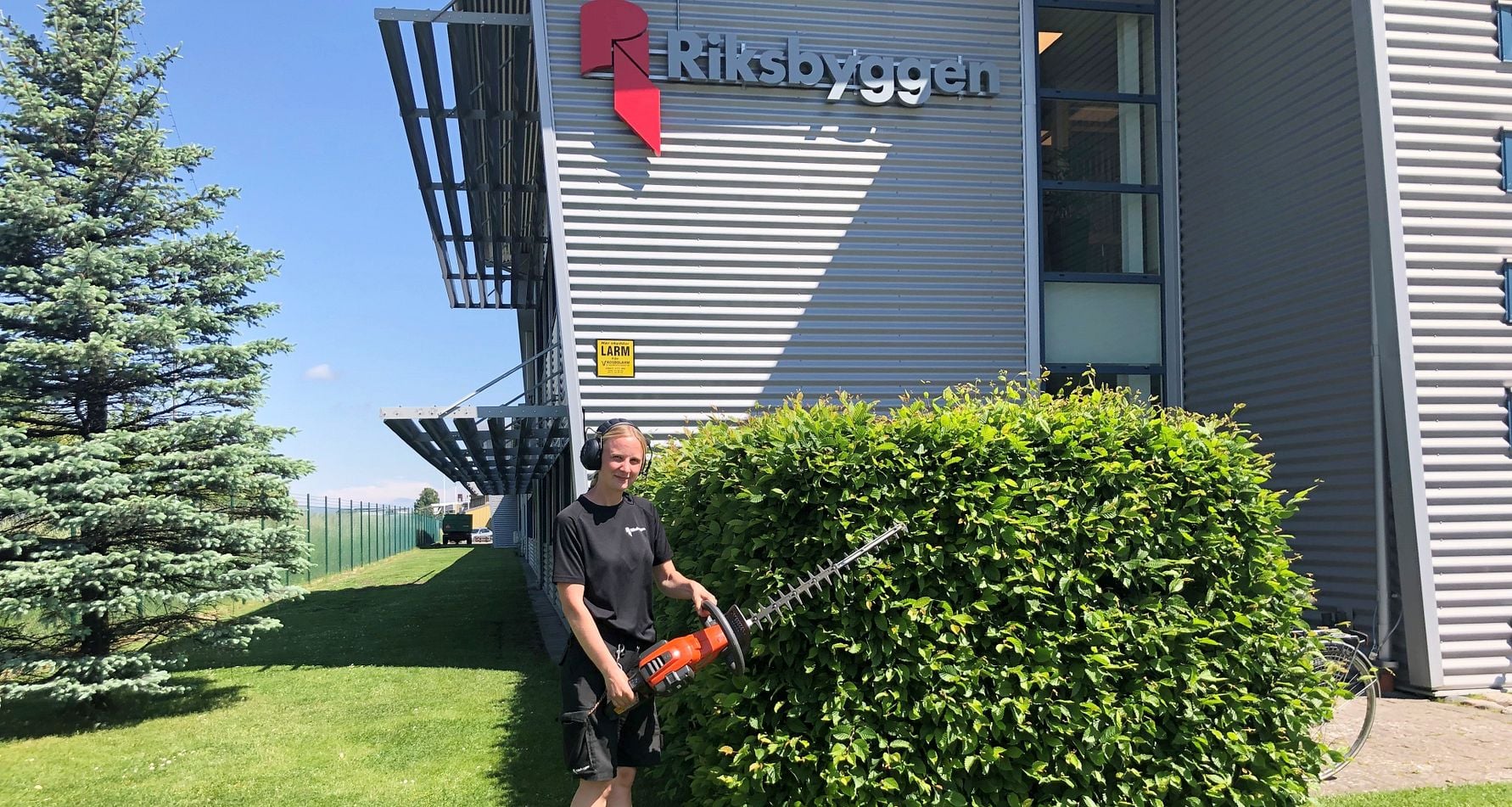 Man klädd i svart, håller en häcksax framför en grön buske vid en byggnad med "Riksbyggen"-skylt
