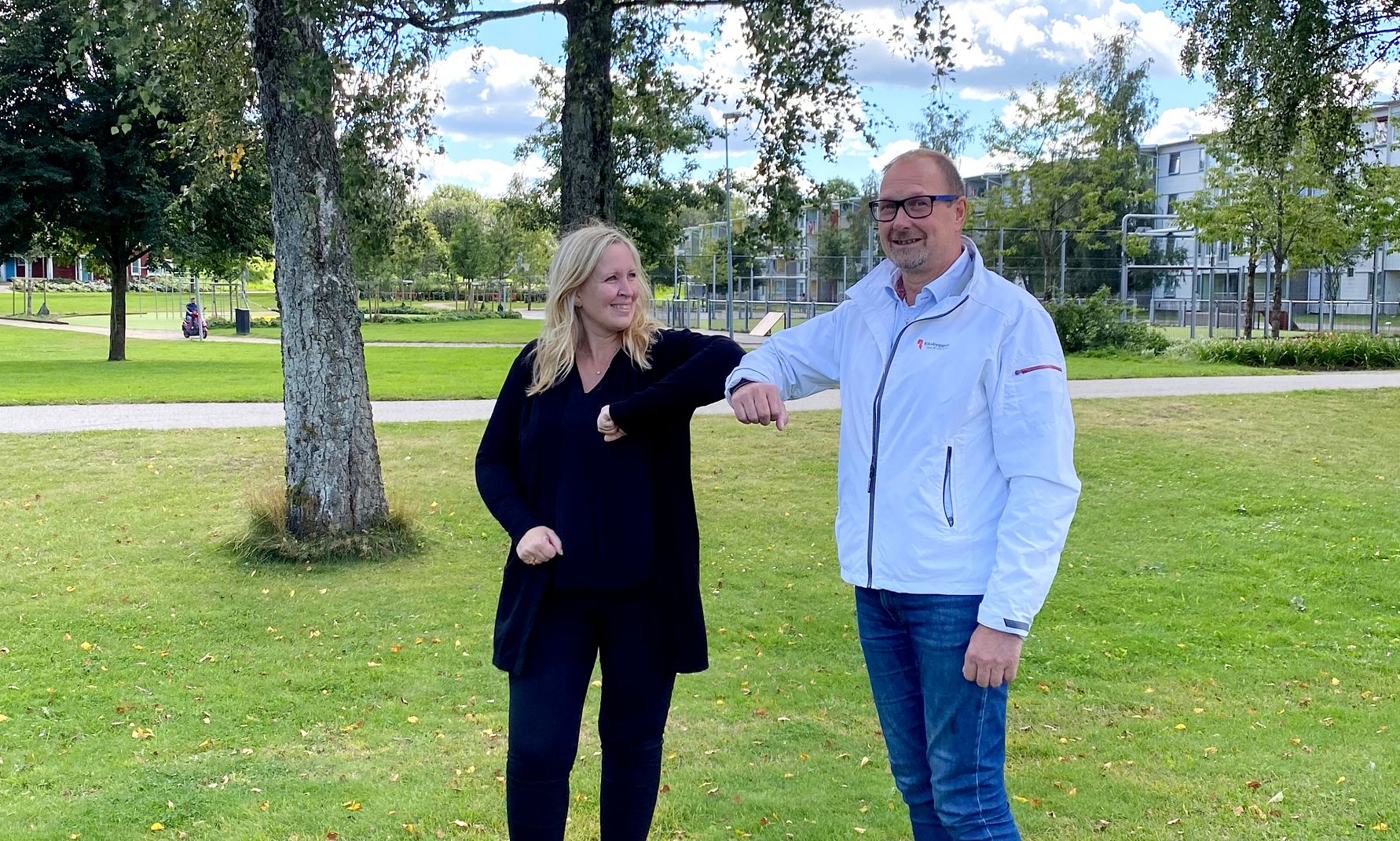 Två personer hälsar på varandra med armbågarna i en park med grönt gräs, träd och blå himmel i bakgrunden
