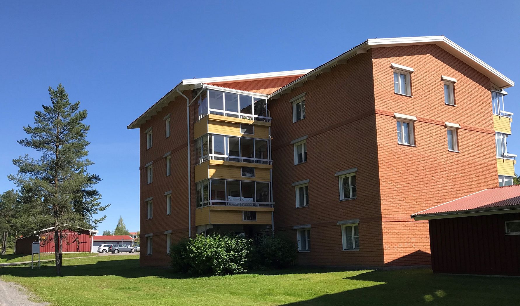 Fyravåningshus i rött tegel med balkonger, omgiven av grön gräsmatta och träd, under en klar blå himmel
