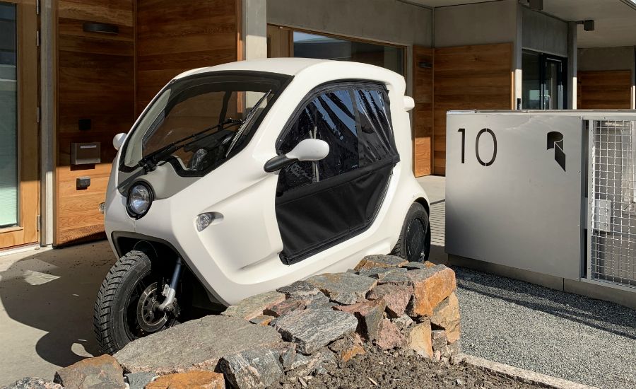 Elbil med tre hjul parkerad framför en modern byggnad med träpanel och stenmur