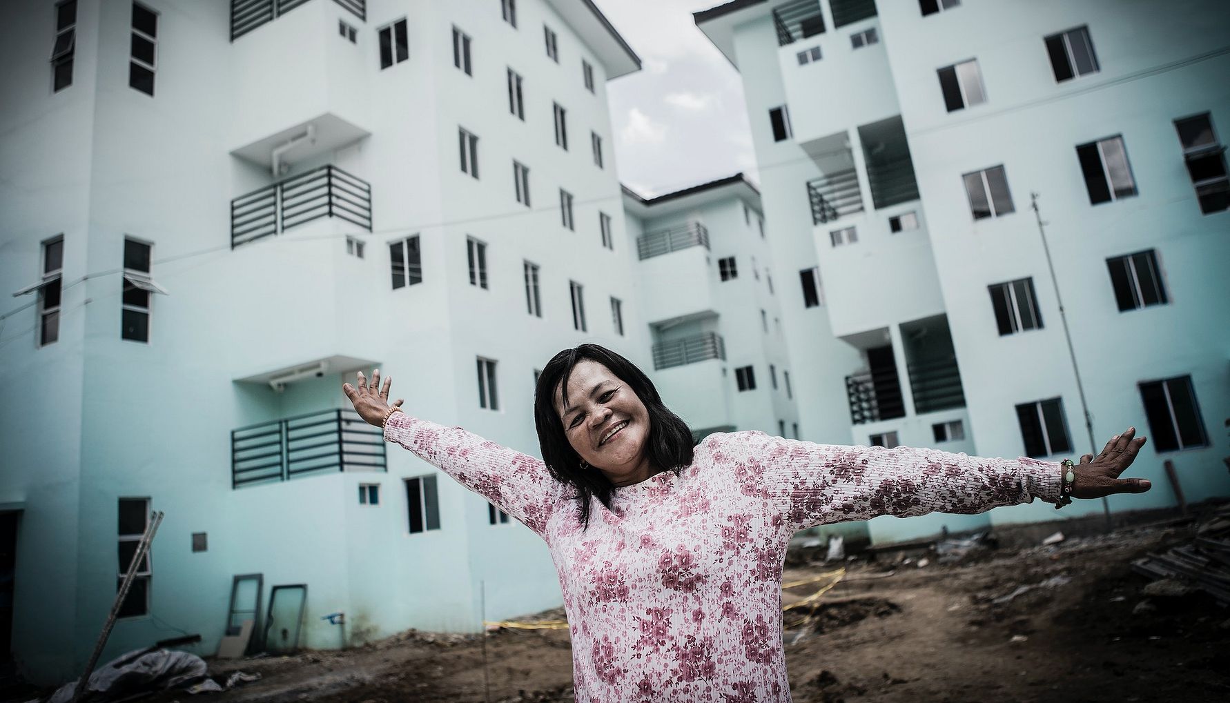 Kvinna i blommig tröja står med utsträckta armar framför ljusblå flerbostadshus under byggnation