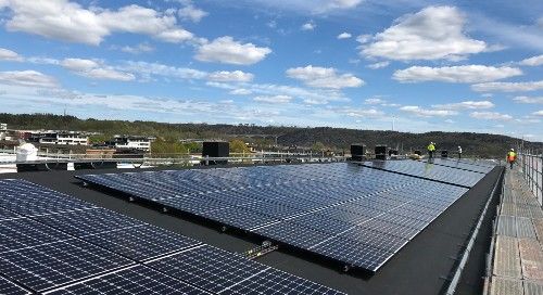 Solpaneler installerade på tak med utsikt över en stad och skog i bakgrunden under en klar blå himmel med moln