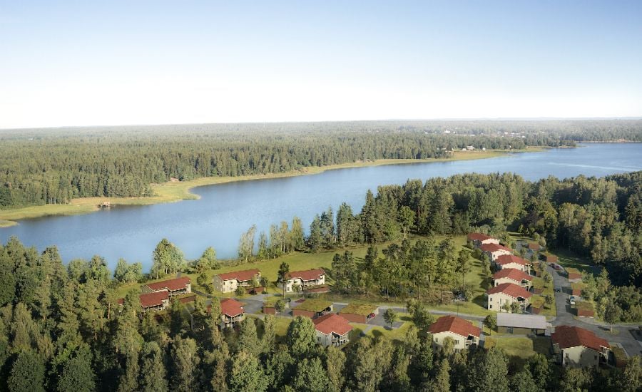 Flygbild av ett bostadsområde med röda tak, omgiven av skog och beläget vid en sjö