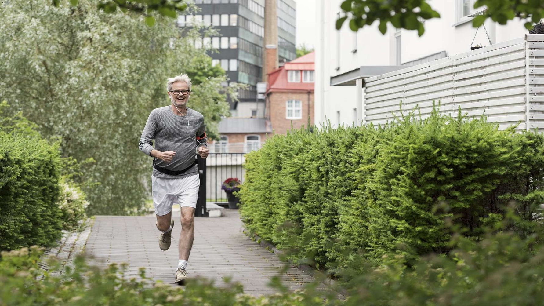 Äldre man joggar på en stenlagd gångväg omgiven av gröna buskar och träd, med stadsmiljö och byggnader i bakgrunden