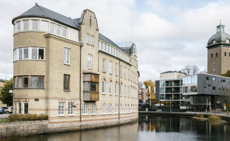 Byggnad med gul fasad vid en kanal, med en modern byggnad och en kyrka i bakgrunden under molnig himmel