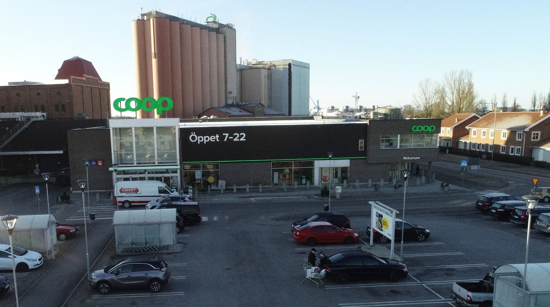 Bild av en Coop-butik med parkeringsplats i förgrunden och ett stort lagerbygge i bakgrunden. Texten "Öppet 7-22" syns på butikens fasad