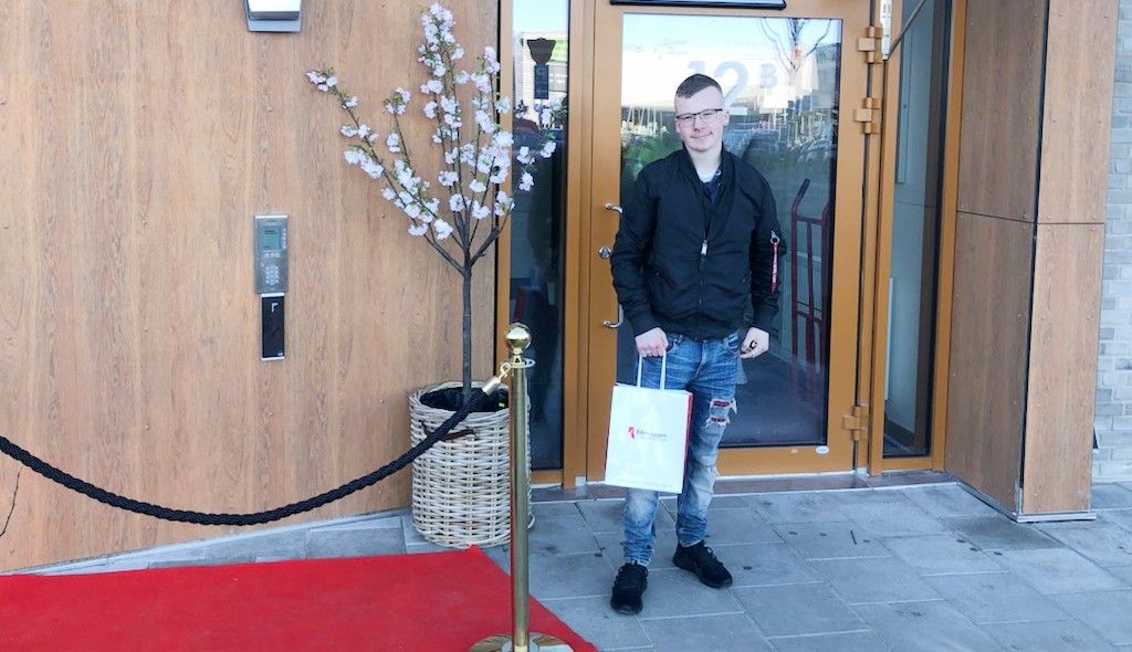 En person står utanför en entré med träpanel och glasdörrar, hållandes en vit papperspåse. Bredvid finns en blommande dekorativt träd i en flätad korg och ett rött rep markerar en gång på en röd matta