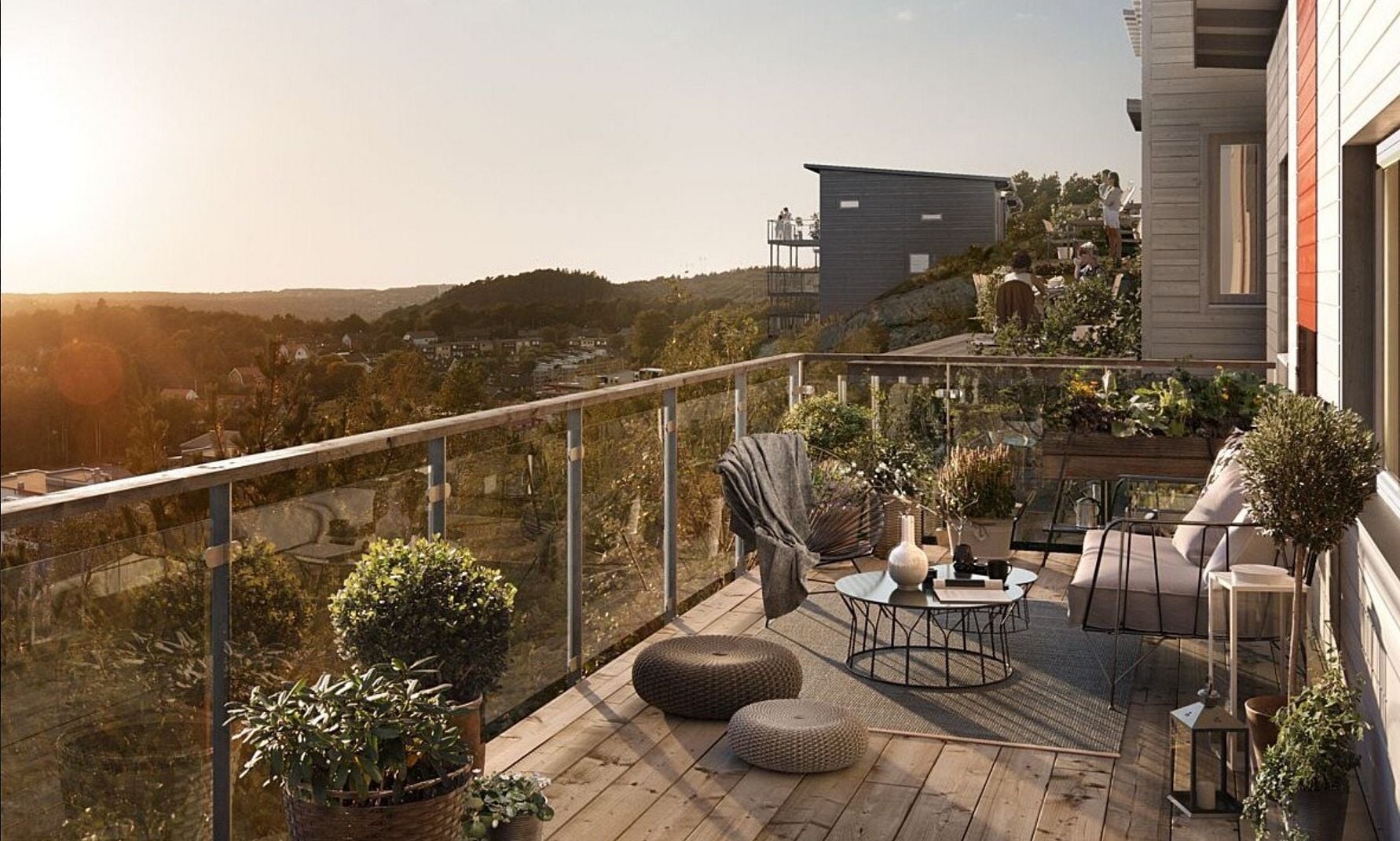 En solig terrass med trämöbler, glasräcke och utsikt över ett naturskönt område med hus och träd i bakgrunden