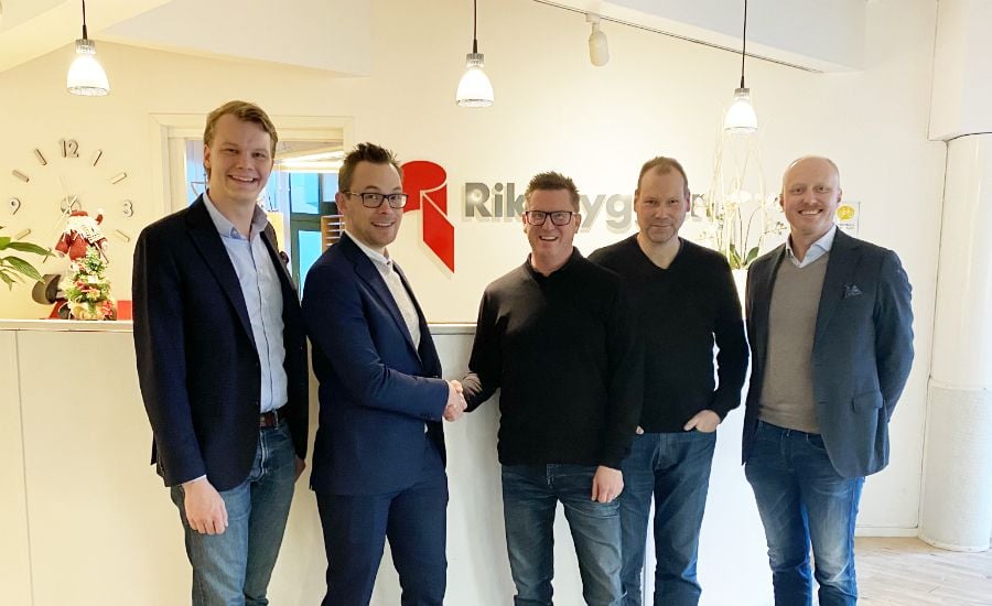 Fem personer står framför en reception med en skylt som visar "Riksbyggen". Två av personerna skakar hand