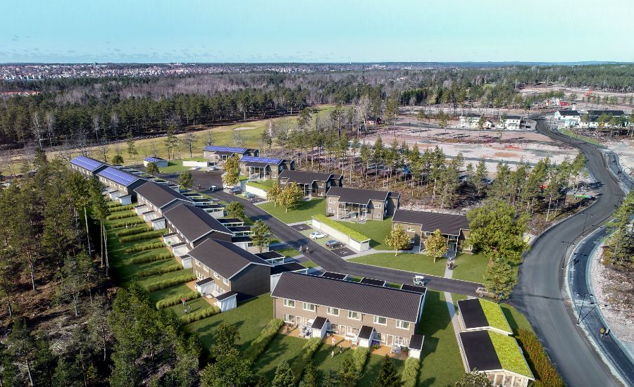 Luftvy av ett bostadsområde med moderna hus omgivna av skog och öppna ytor, inkluderar vägar och parkeringsområden