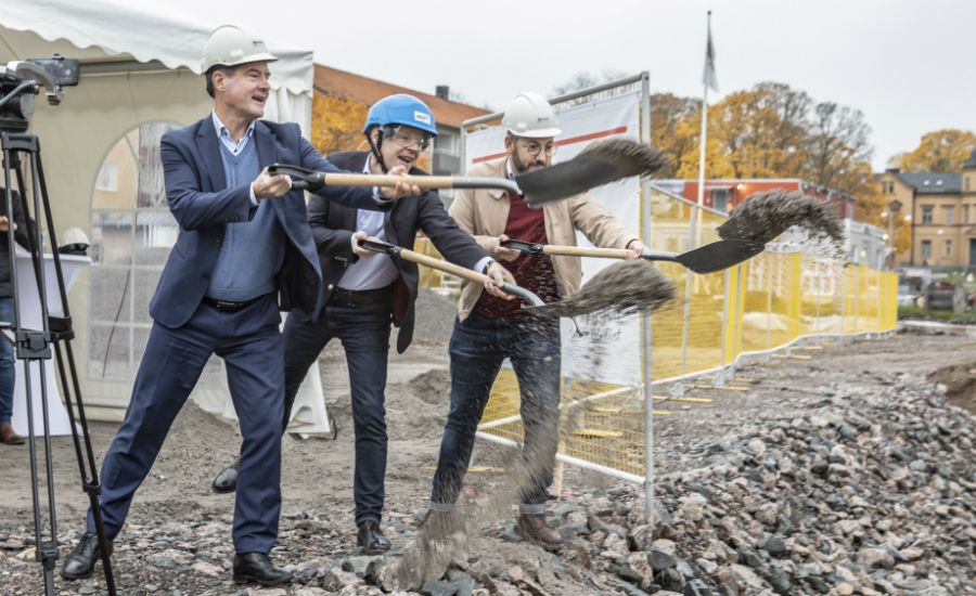 Personer med bygghjälmar använder spadar för att symboliskt skyffla jord vid en byggarbetsplats, med tält och byggnadsstängsel i bakgrunden