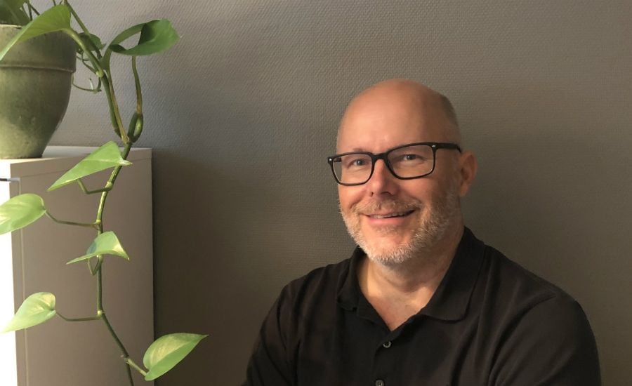 Man i svart pikétröja sitter bredvid en grön växt i en grå miljö