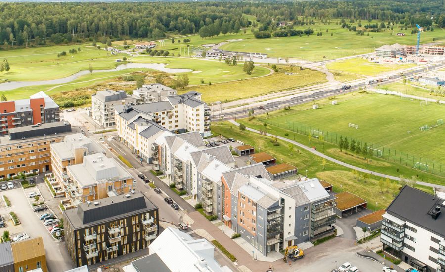 Flygvy över ett bostadsområde med moderna byggnader, omgivande grönområden och en fotbollsplan